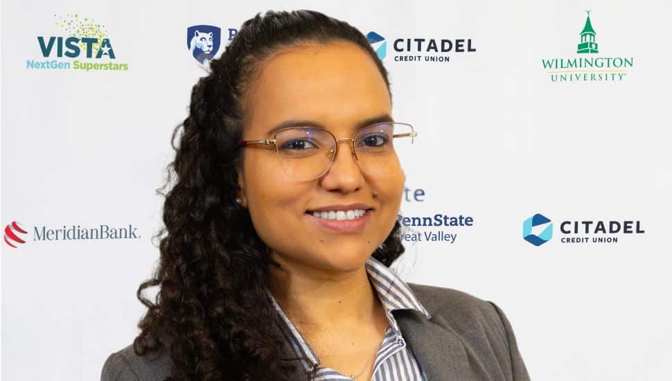 Headshot of Nathalia Nascimento. She has dark curly long hair, wears a gray blazer and glasses, and is smiling.