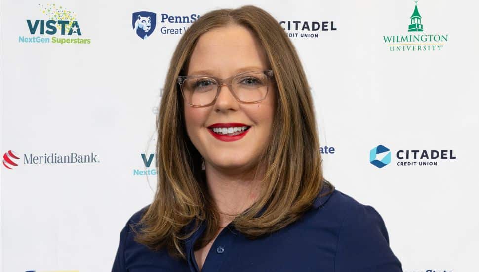Tess Protesto stands in front of the VNS2025 banner and wears a navy colored shirt, red lipstick, glasses, long brown hair, and is smiling.