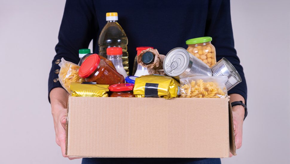 A person in a black shirt holds a box filled with dry and canned goods.