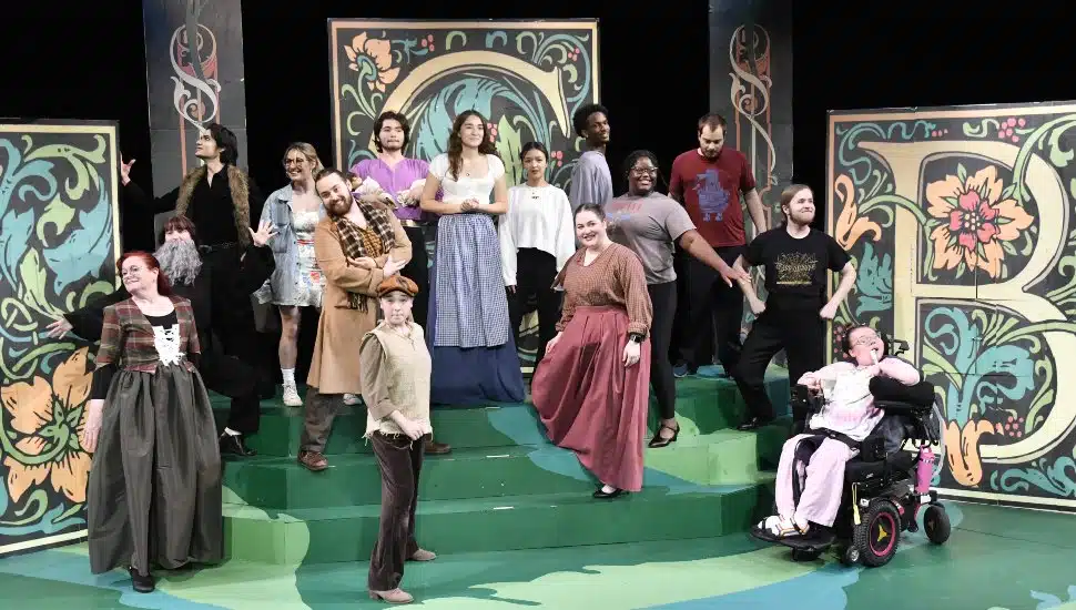 DCCC cast members of "Into the Woods" on stage in full costume. The set is woodsy and earthy in nature, with hues of gold, green, and brown.