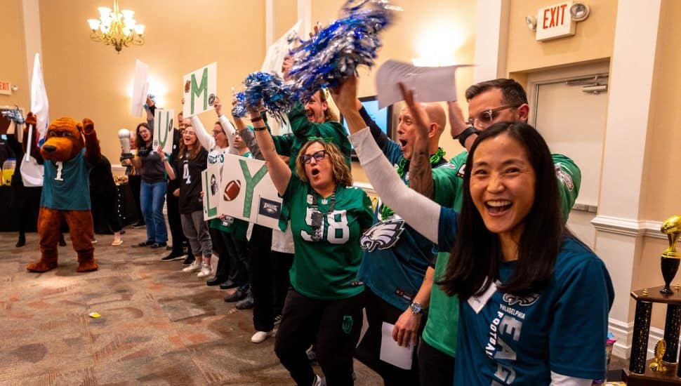 Employees of the YMCA of Greater Brandywine enthusiastically cheer in matching Philadelphia Eagles gear during an indoor celebration, holding up signs and pom-poms. A person in a bear mascot costume and festive trophies are visible in the background, creating a lively and team-spirited atmosphere.