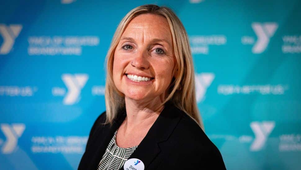 Diana Beeson, newly appointed Associate Vice President of Membership at the YMCA of Greater Brandywine, smiles confidently while wearing a black blazer and name badge. She stands in front of a teal YMCA-branded backdrop featuring multiple Y logos and the words "YMCA of Greater Brandywine."