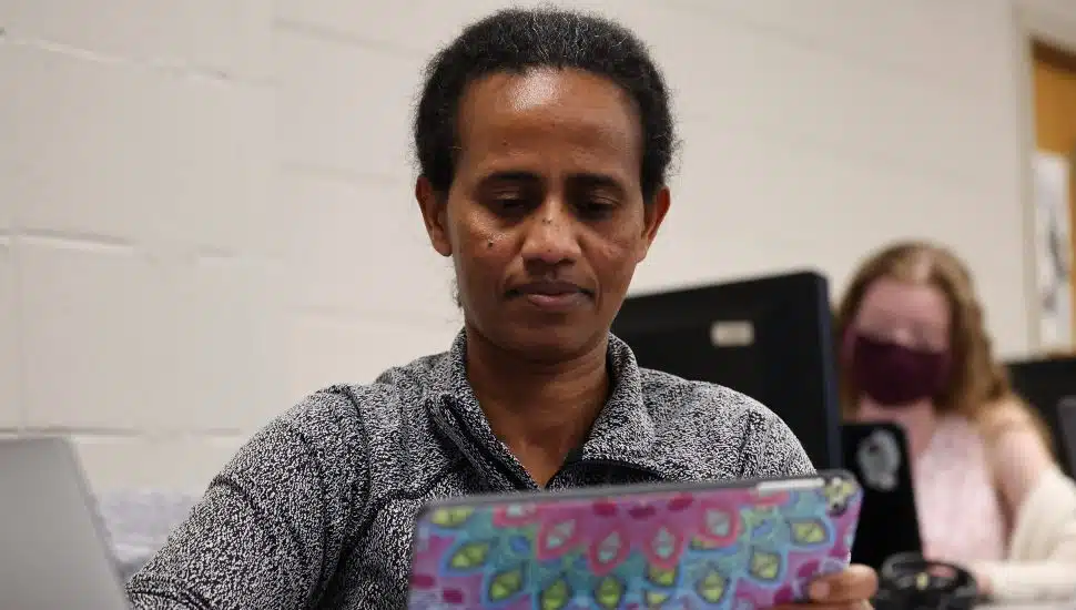 Adult student focused on a colorful tablet during a class session, with another student working at a computer in the background.