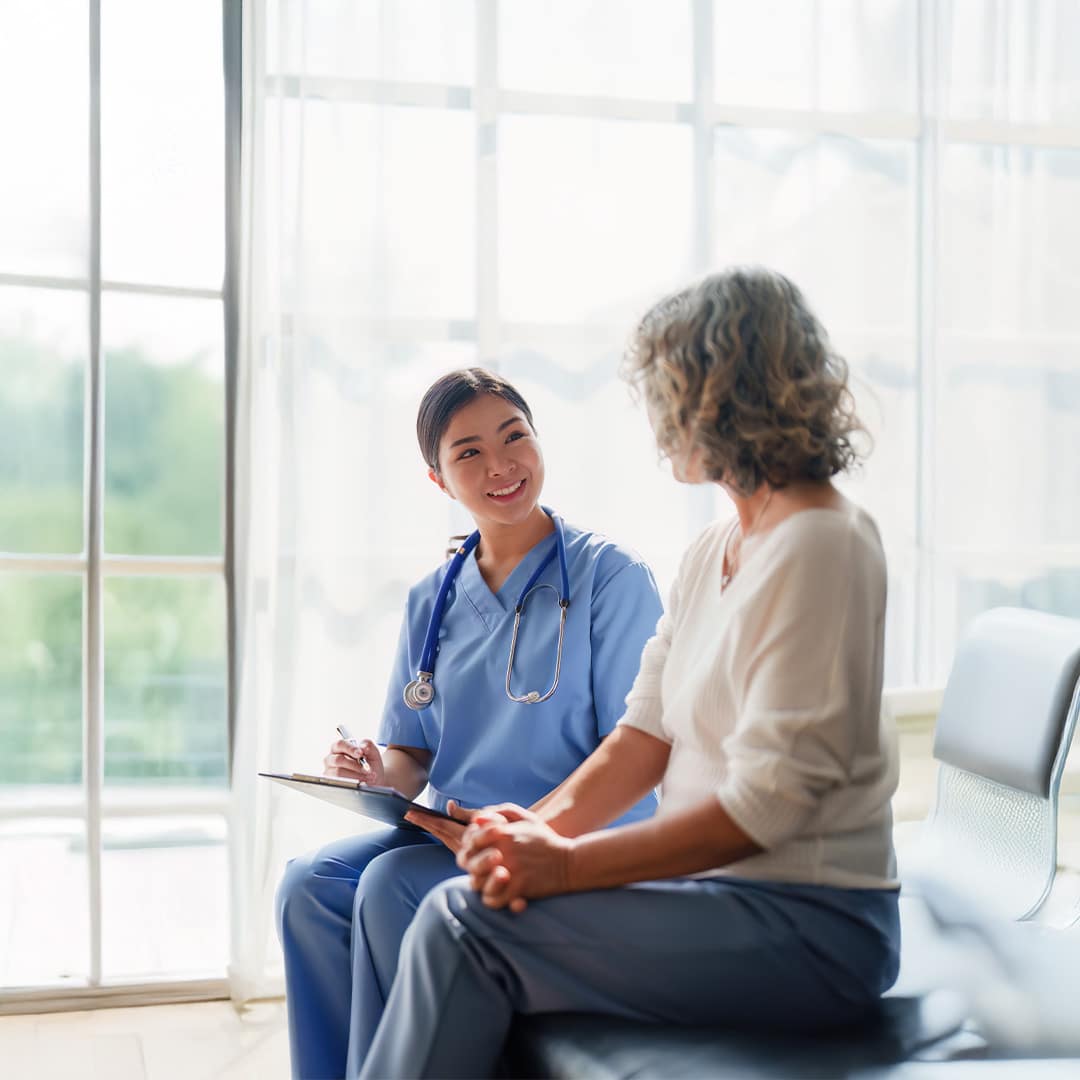 A nurse practitioner sits with a clipboard talking to a woman in a medical room/waiting room area.