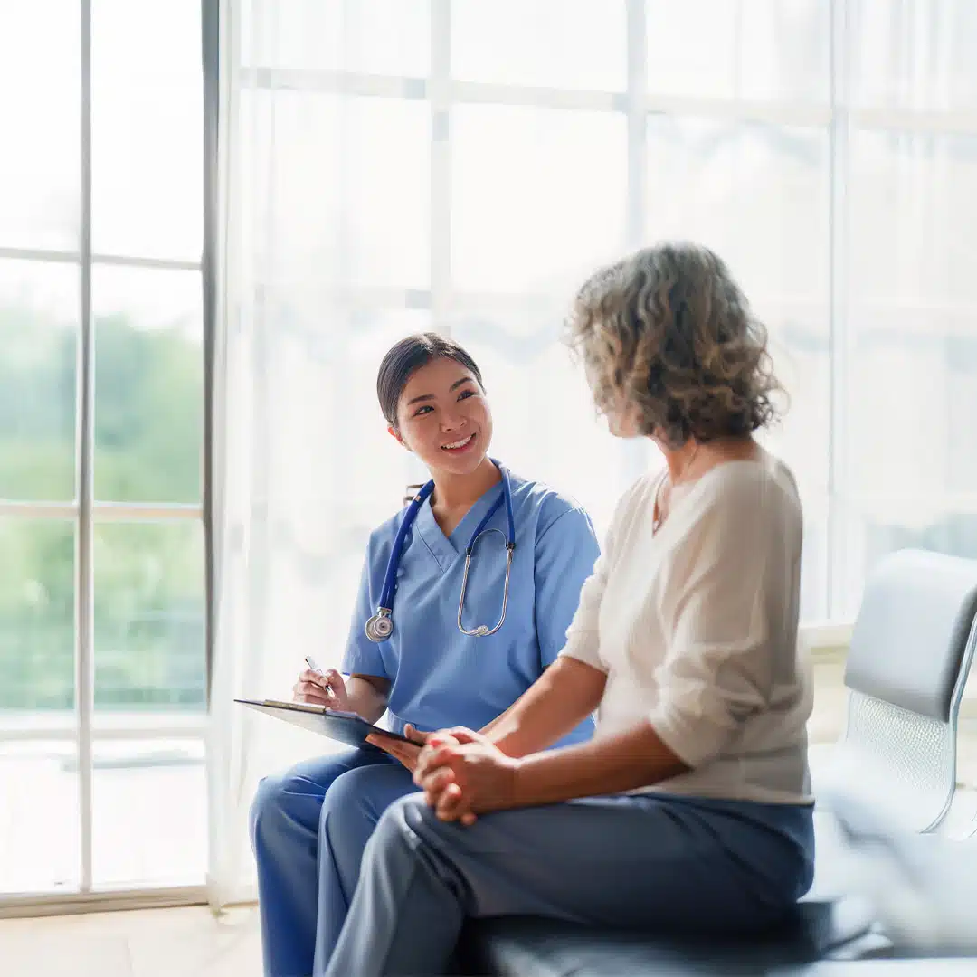 A nurse practitioner sits with a clipboard talking to a woman in a medical room/waiting room area.