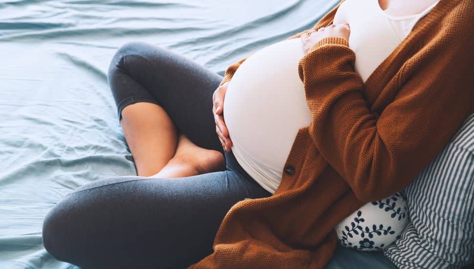 Beautiful pregnant woman sitting at bed and holds hands on belly in bedroom at home. Young mother waiting of a baby. Pregnancy, maternity and expectation image. Close-up, indoors.