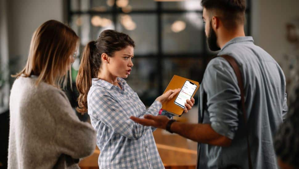 Three people in a discussion, with one woman showing concern while holding a smartphone displaying a credit score app, as she talks to a man and another woman in a casual indoor setting.