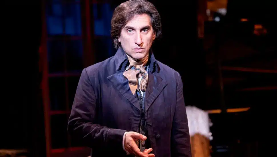 Hershey Felder on stage dressed as Fryderyk Chopin, standing center stage in a warmly lit Parisian salon set.