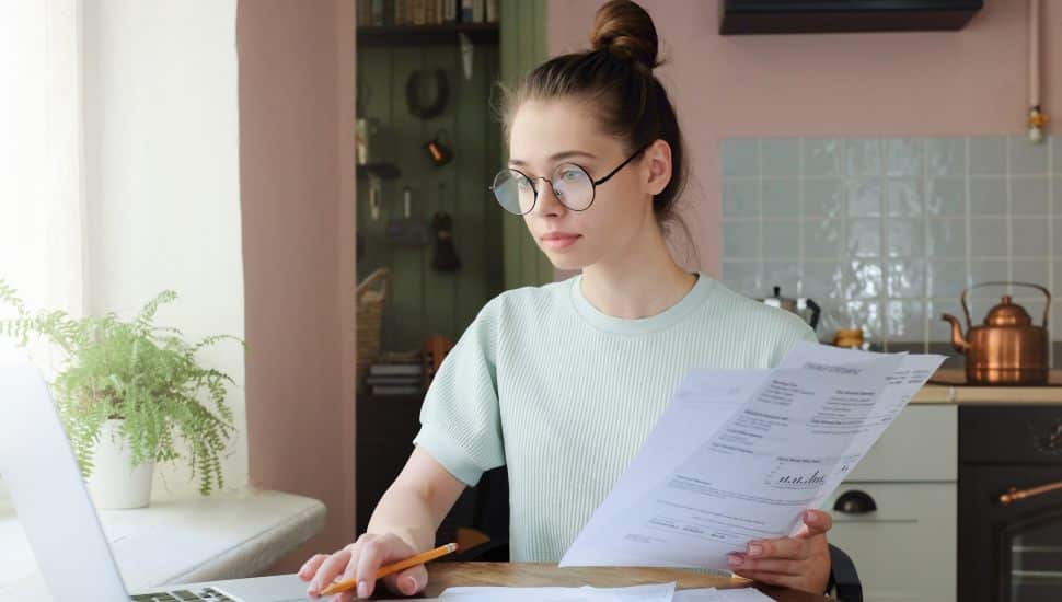 Young woman managing budget, sitting at kitchen table with open laptop, documents and calculator, using touchpad, making notes with pencil.