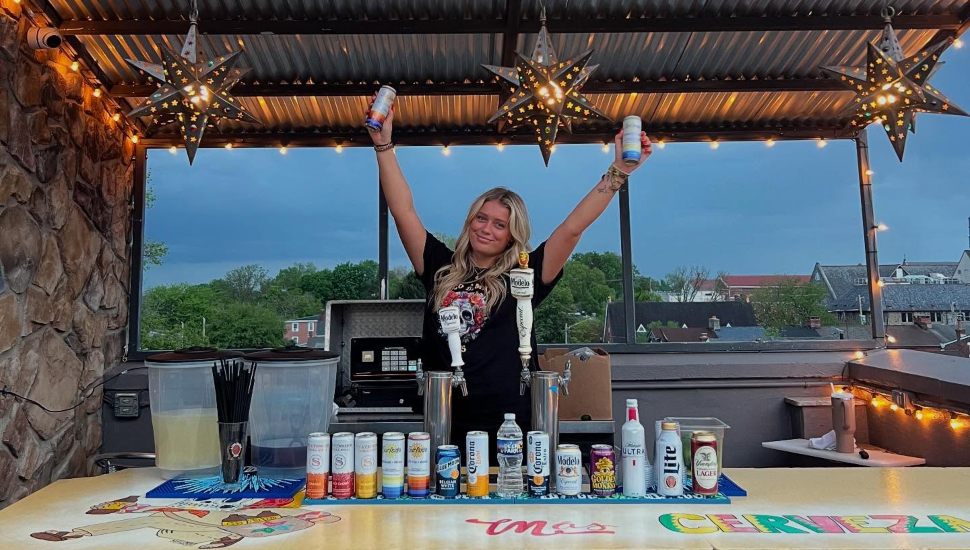 Blonde bartender holds seltzers in air behind bar on rooftop of Mas.