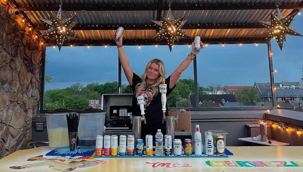 Blonde bartender holds seltzers in air behind bar on rooftop of Mas.