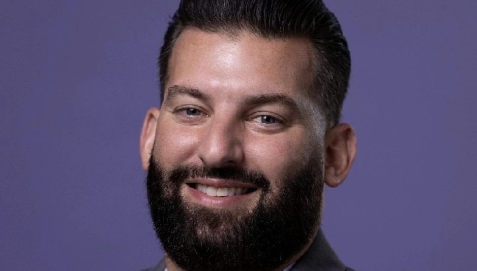 Headshot of Conte against a purple background: He is smiling, has a dark beard and hair, and wears a jacket and tie.