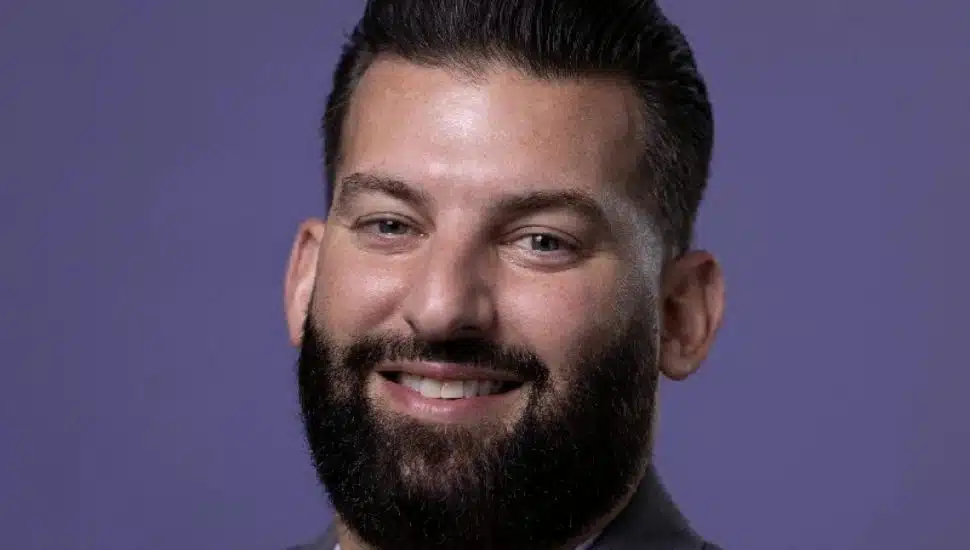 Headshot of Conte against a purple background: He is smiling, has a dark beard and hair, and wears a jacket and tie.