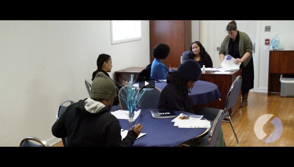 A group of volunteers and adult learners gather at a few tables. There are stacks of paper and everyone is focused on learning.