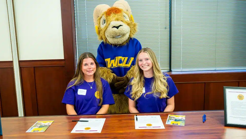 Two young women sit at a table smiling, each wearing a purple shirt with a gold logo. In front of them are official-looking documents and pens. Behind them stands a person in a ram mascot costume wearing a blue and gold “WCU” shirt, smiling for the photo. The setting appears to be an indoor office or school environment with wooden paneling and large windows with blinds.