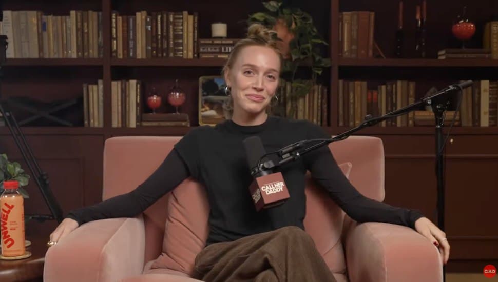 Alex Cooper sitting in a chair with microphone in front of her and bookcase behind.