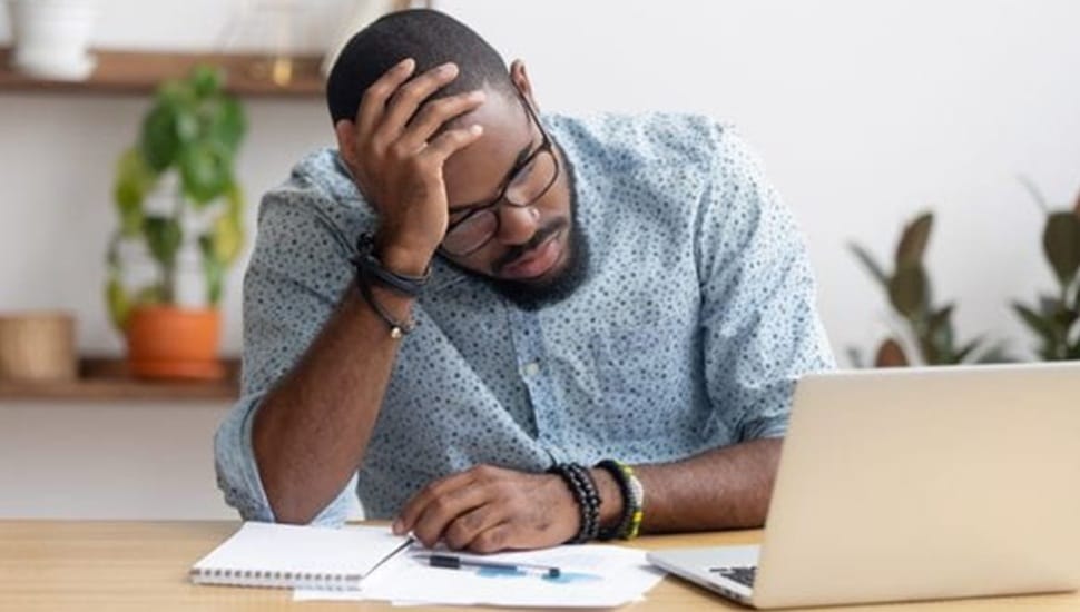 Young black male with beard and glasses sits frustrated a laptop computer.