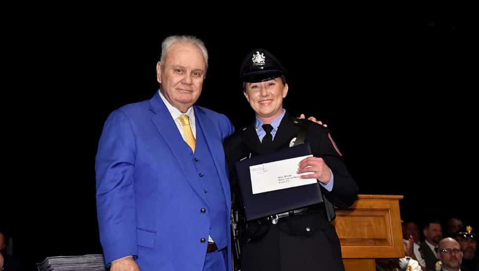MPA Director William Davis with Cadet Ariel Brown of Philadelphia. Police academy graduate in uniform smiles while holding a certificate, standing next to a man in a blue suit during a graduation ceremony.          
