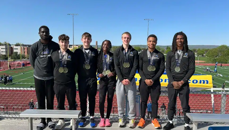 Group photo of seven DCCC Phantoms Track and Field athletes standing on bleachers in front of a track and field stadium under a clear blue sky. From left to right: Dennis Okane-Nketia (relay alternate) wearing a dark team sweatshirt and black pants, Aidan Heppard in black athletic gear with two medals, Jimmy Love in black athletic gear with multiple medals, Fatumata Kaba in black athletic gear with multiple medals and holding a baton, Daniel McLaughlin in black athletic jacket and gray sweatpants with medals, Fred 'Tre' McCray in black athletic gear with multiple medals, and Daishon Billops in black athletic gear with medals. The athletes smile proudly after excelling at Nationals. DCCC banner and sports field visible in the background.