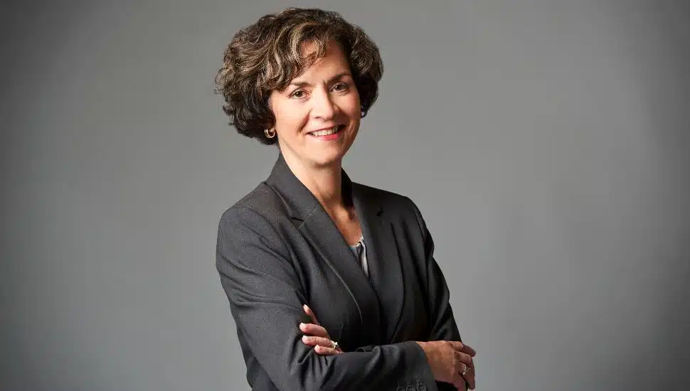 Dr. Marta Yera Cronin, President of Delaware County Community College, poses confidently in a professional dark gray suit against a plain gray background, smiling with arms crossed.