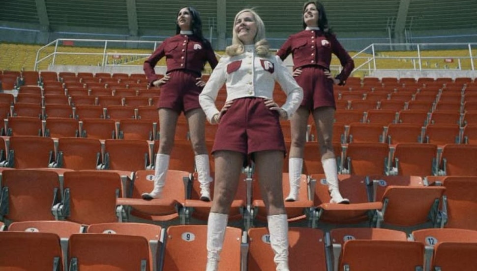 Three women from "Hot Pants Patrol" standing on chairs in baseball stadium.