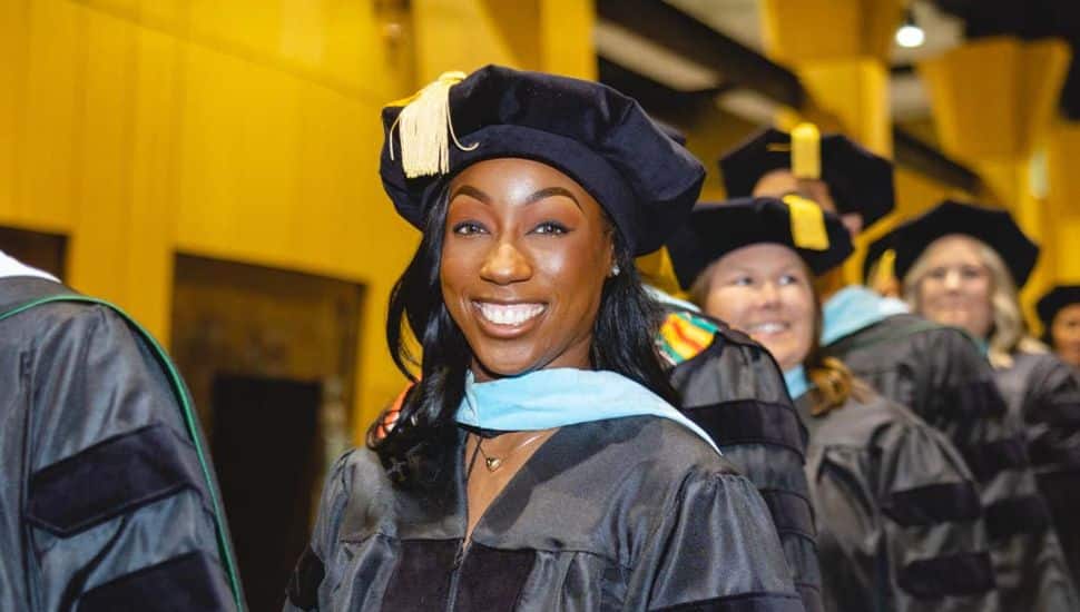 Smiling woman in doctoral regalia at Wilmington University graduation ceremony, surrounded by fellow graduates in academic gowns and caps.