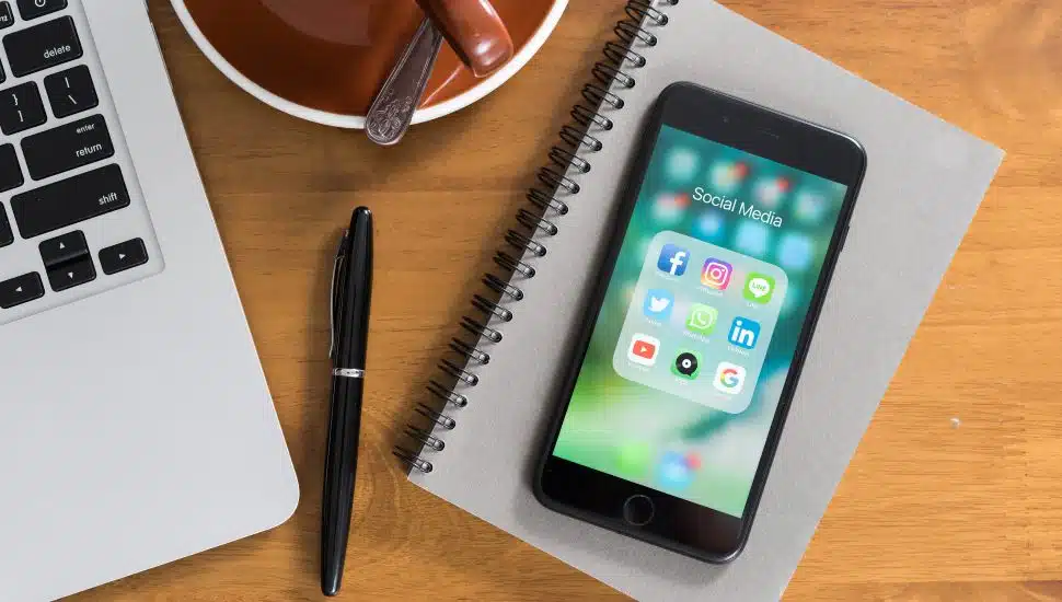 A phone showing social media icons, pictured next to a cup of coffee, laptop and notebook and pen. Concept of research, networking, career advancement.