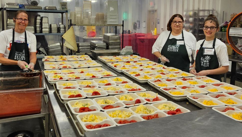 Three Chester County Food Bank volunteers smiling while preparing and packaging rows of colorful prepared meals in a commercial kitchen.