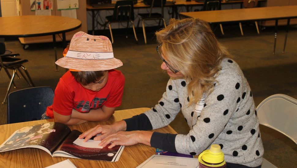 Adult volunteer helps a young boy read a picture book during a classroom literacy session. The boy wears a handmade paper hat, and they are seated at a table with books and school supplies.