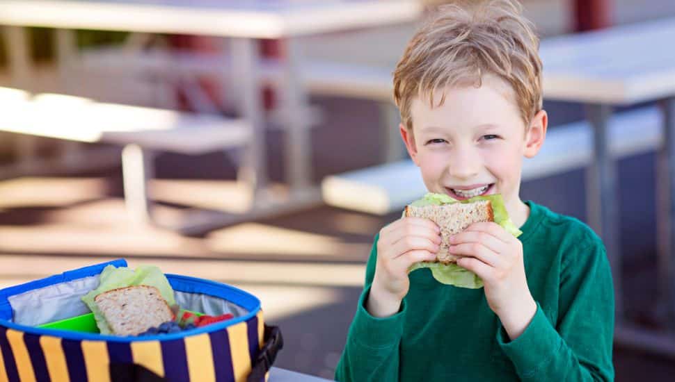 Smiling young boy in a green shirt eating a sandwich outdoors at a picnic table, with an open striped lunchbox containing more food items beside him.