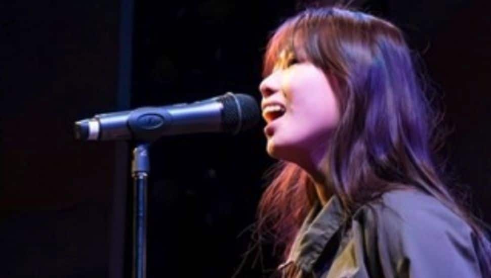 A young performer sings into a microphone on stage under theatrical lighting, wearing a green jumpsuit during a musical theatre showcase.