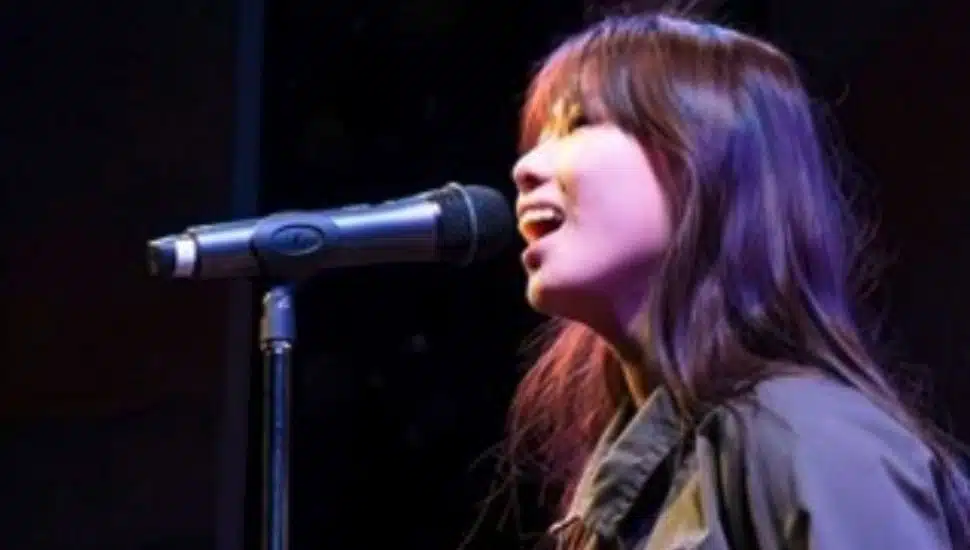 A young performer sings into a microphone on stage under theatrical lighting, wearing a green jumpsuit during a musical theatre showcase.