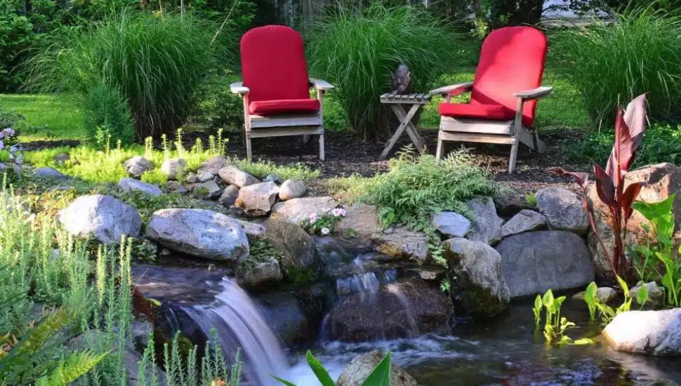Garden with a small waterfall surrounded by rocks and plants, with two wooden chairs with red cushions and a small table in the background.