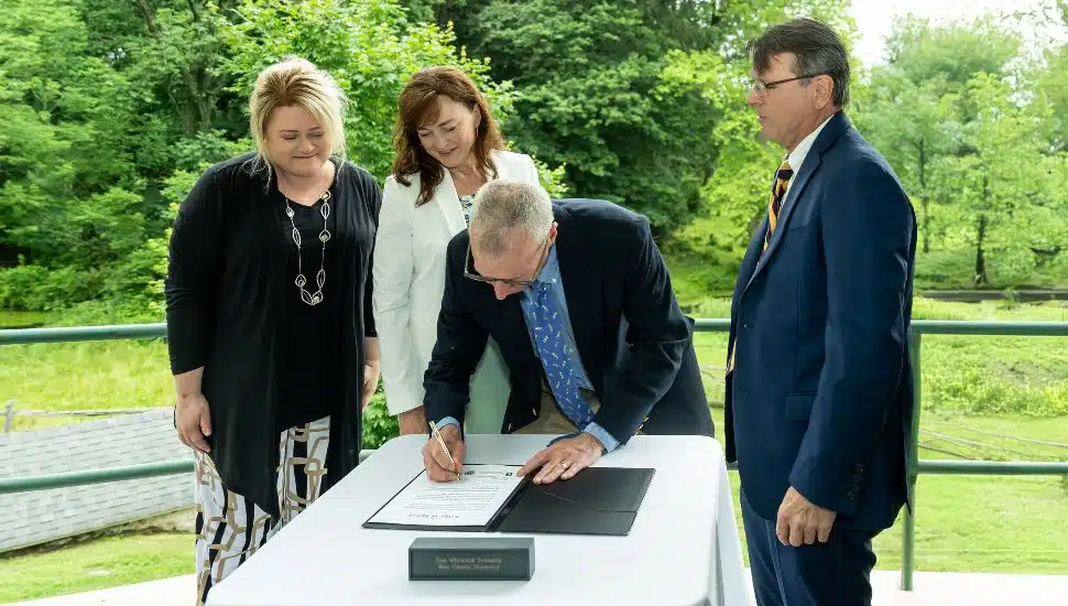 West Chester University and East Whiteland Township held a formal signing ceremony to celebrate their historic partnership in preserving a 52-acre farm that was gifted by an anonymous donor. Pictured (L to R) are West Chester University President Laurie Bernotsky, Executive Director of the West Chester University Foundation Deb Cornelius, East Whiteland Township Manager Steve Brown (signing), and Chairman of the East Whiteland Township Board of Supervisors Scott Lambert. A man in a suit signs a document on a table outdoors, while three other professionally dressed individuals—two women and one man—stand beside him, observing. The scene is set against a backdrop of lush green trees, suggesting a formal signing event in a natural setting.