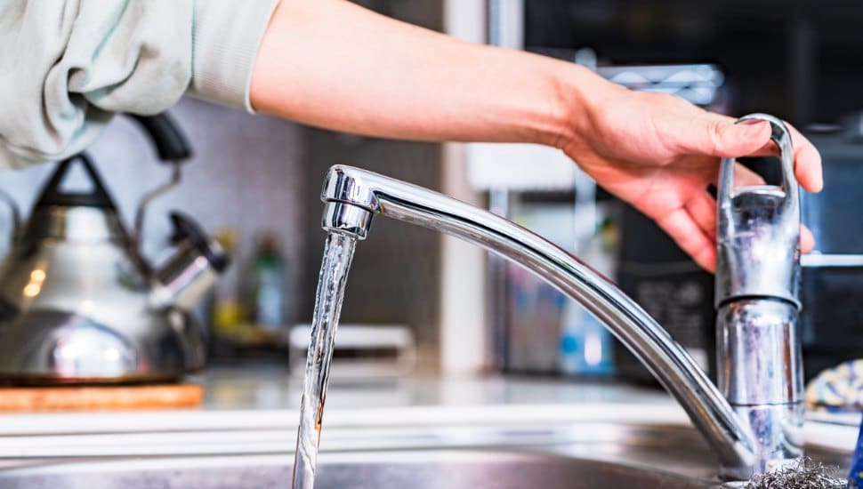 A person turning on a kitchen faucet with water flowing, in a bright home setting with a kettle in the background.
