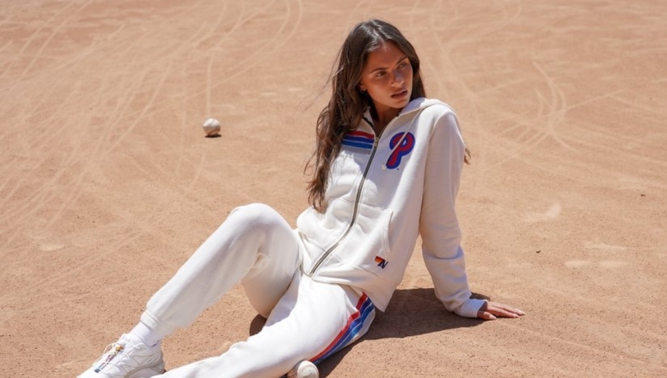 Woman wearing hoodie and sweatpants designed for the Aviator Nation x Philadelphia Phillies apparel collection with baseball behind her.