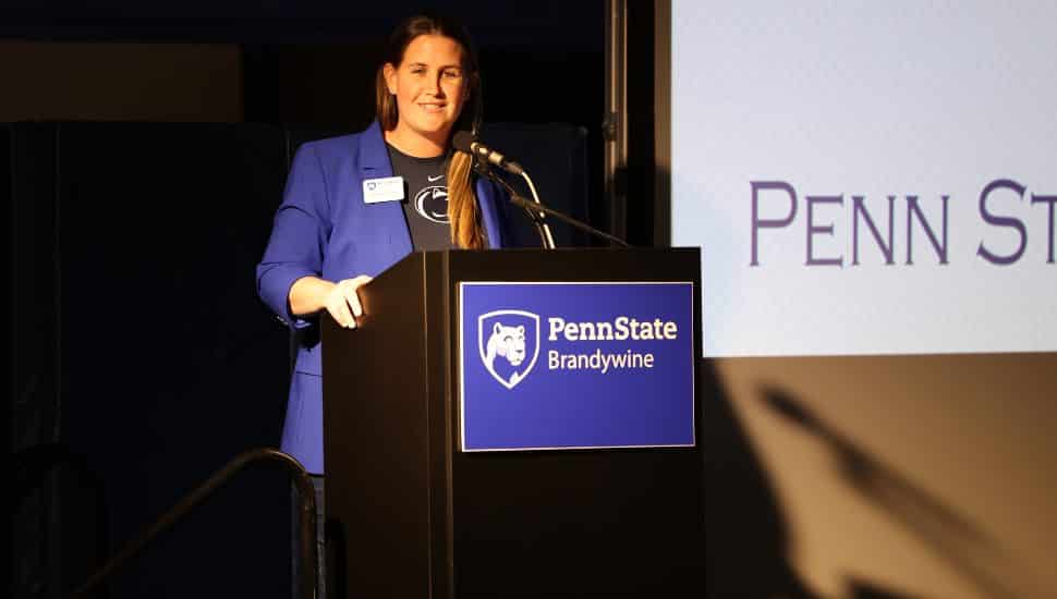Bobbi Caprice stands at a podium labeled 'Penn State Brandywine,' speaking into a microphone. She is wearing a blue blazer over a Penn State shirt, smiling while addressing an audience at an indoor event.