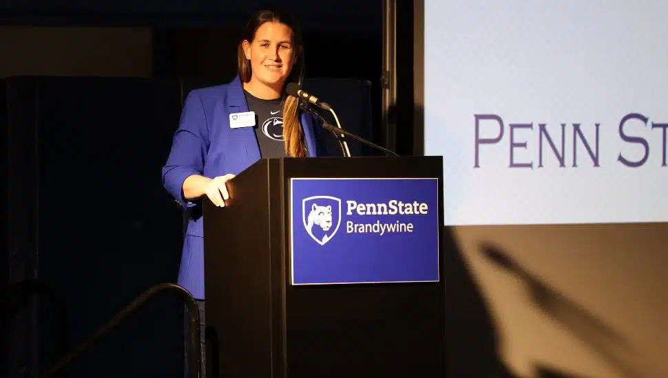 Bobbi Caprice stands at a podium labeled 'Penn State Brandywine,' speaking into a microphone. She is wearing a blue blazer over a Penn State shirt, smiling while addressing an audience at an indoor event.