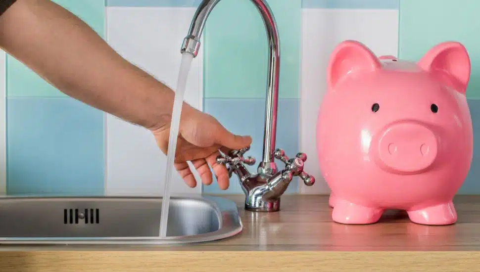 Person turning off a kitchen faucet with a pink piggy bank nearby, symbolizing water conservation and saving money on utility bills.