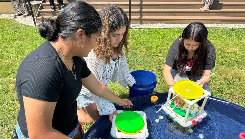 Three high school students participating in a hands-on STEM activity at Washington College, testing floating data buoys in a water-filled basin during a summer immersion program.