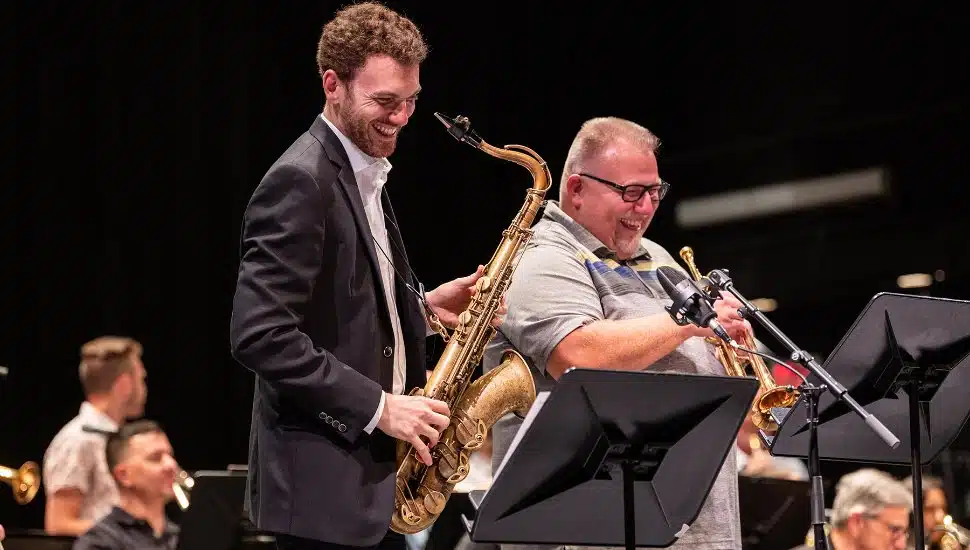 Two musicians, one playing a saxophone and the other a trumpet, smile and share a light moment on stage during a live jazz performance, with other band members visible in the background.