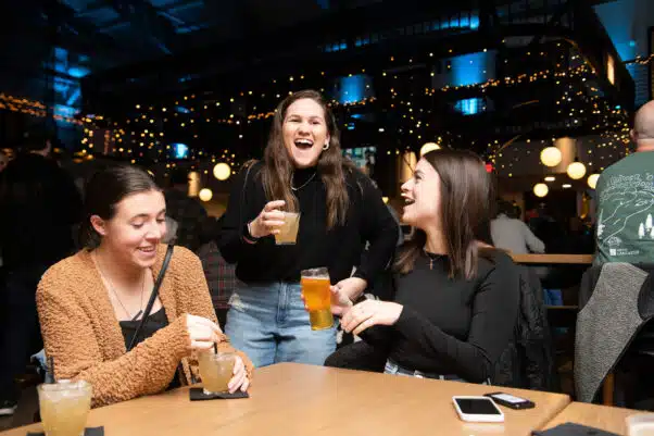 Three young women enjoy drinks and laughter at Southern Market in Lancaster, seated at a wooden table under warm string lights and a festive indoor atmosphere.