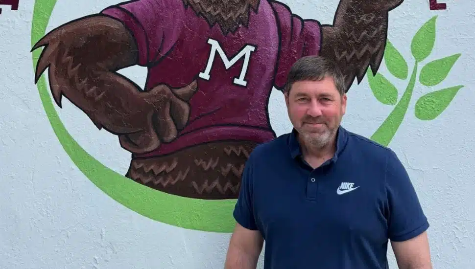 Middle-aged white male in blue polo shirt poses for picture in front of painting of school mascot.