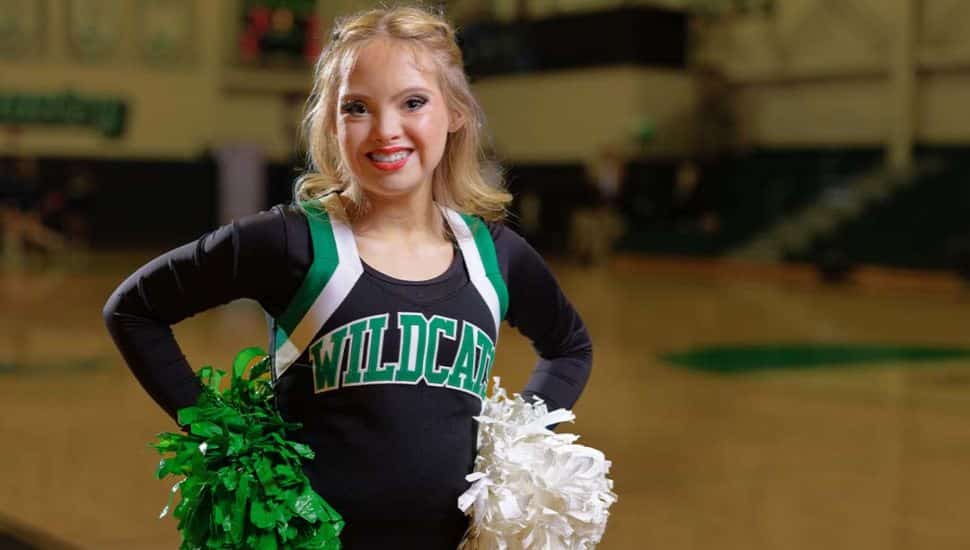 Kayla Kosmalski, a Wilmington University cheerleader with Down syndrome, smiles confidently in her uniform holding green and white pom-poms on a basketball court.