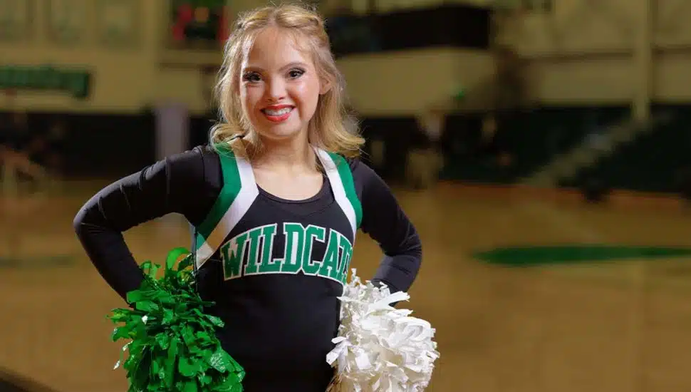 Kayla Kosmalski, a Wilmington University cheerleader with Down syndrome, smiles confidently in her uniform holding green and white pom-poms on a basketball court.