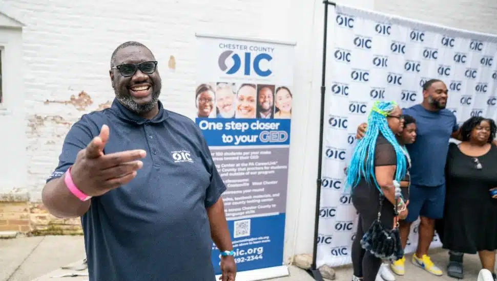A man in a Chester County OIC polo shirt smiles and gestures toward the camera while others pose with Eagles player Brandon Graham in front of an OIC backdrop at the 2025 Block Party.
