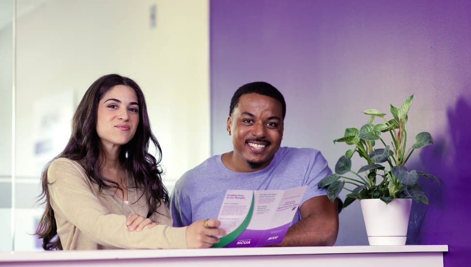 Two smiling young adults, a woman and a man, sit together at a counter holding a brochure in a bright, modern space with a purple wall and a potted plant beside them — conveying a friendly, welcoming atmosphere, likely in a financial or community service setting.