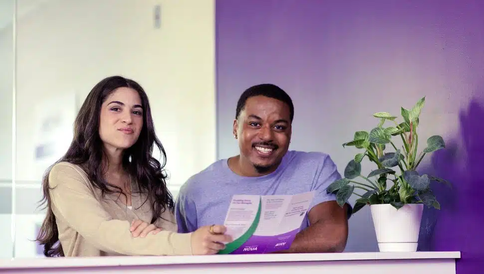 Two smiling young adults, a woman and a man, sit together at a counter holding a brochure in a bright, modern space with a purple wall and a potted plant beside them — conveying a friendly, welcoming atmosphere, likely in a financial or community service setting.
