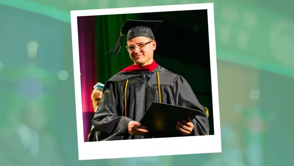 Thomas Burt, dressed in academic regalia, smiles while holding his diploma on stage at Wilmington University’s 2025 commencement ceremony, where he served as the graduate speaker for the College of Technology.