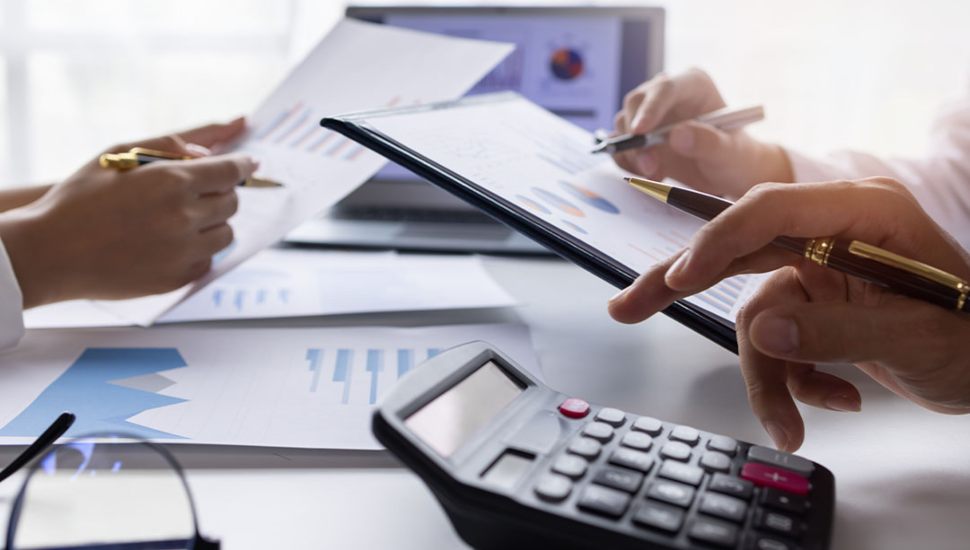 Close-up of hands working with financial charts, a calculator, and documents at a desk, representing accounting or financial analysis work.